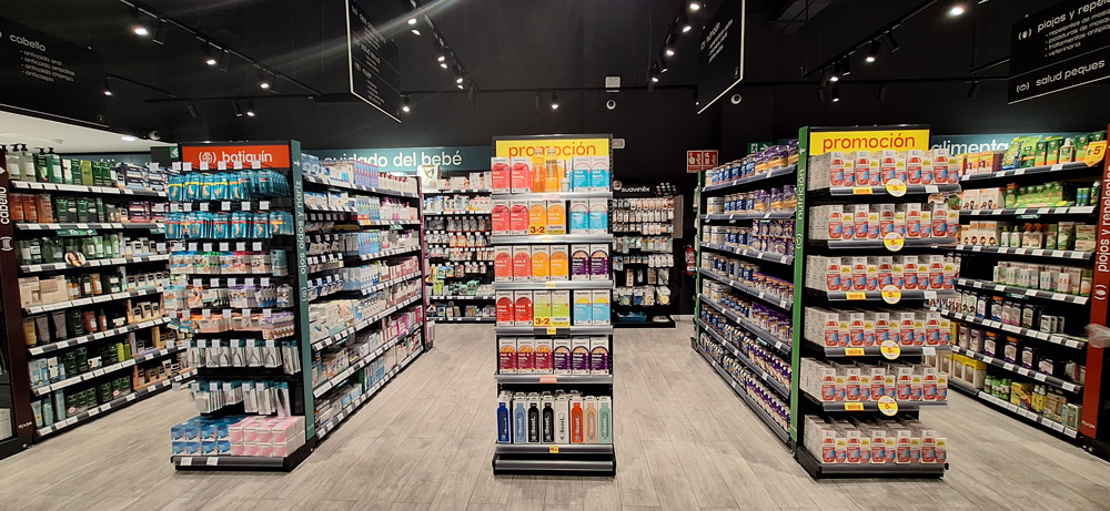 Farmac&eacute;utico en primer plano con los brazos cruzados sonr&iacute;e mientras en el fondo una farmac&eacute;utica ayuda a una clienta con un producto