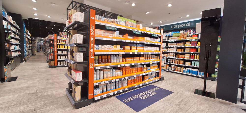 Farmac&eacute;utico en primer plano con los brazos cruzados sonr&iacute;e mientras en el fondo una farmac&eacute;utica ayuda a una clienta con un producto